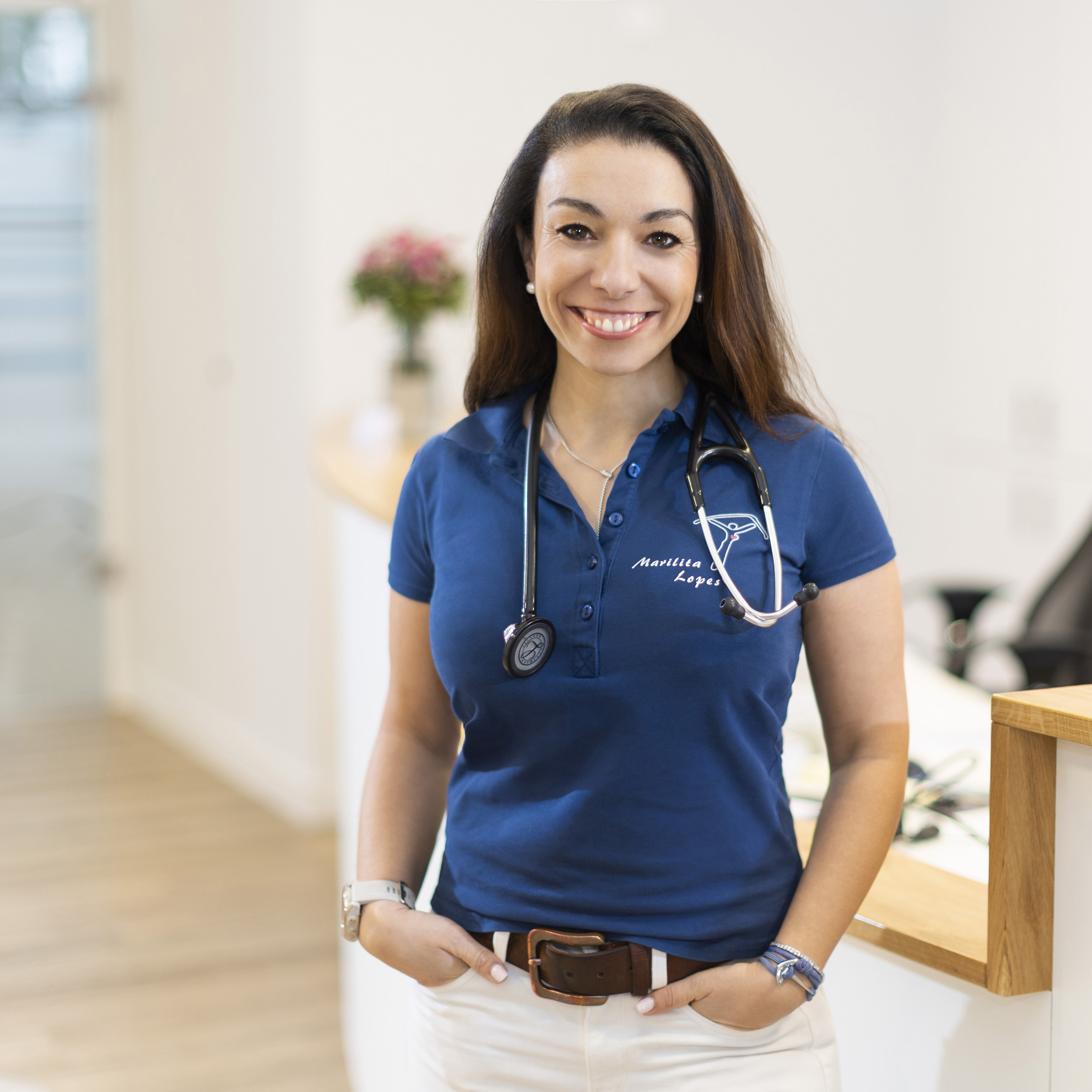 Ärztin in ihrer Praxis, trägt weiße Hose und blaues Shirt mit Stethoskop, lächelnd mit Händen in den Taschen